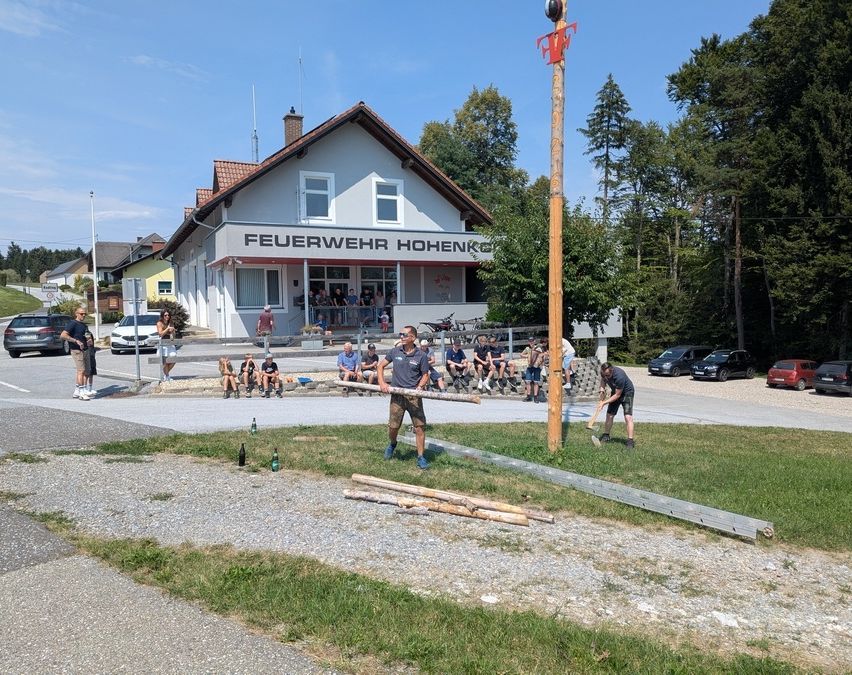 Eine Gruppe von Menschen nimmt an einem Holzhackwettbewerb vor einem Feuerwehrhaus teil. Zuschauer sitzen auf dem Rasen, und zwei Männer hacken mit Äxten Holz.