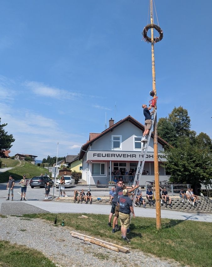 Eine Gruppe von Menschen versammelt sich um einen hohen Holzpfahl mit einem Kranz oben. Ein Mann klettert eine Leiter hinauf, um die Spitze zu erreichen. Im Hintergrund ist ein Gebäude mit dem Schild 'Feuerwehr' zu sehen.