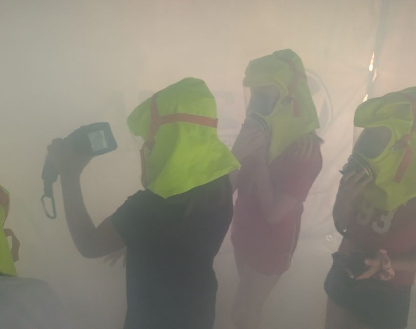 Drei Personen in Schutzkleidung, einschließlich Masken, stehen in einer nebligen Umgebung. Eine Person hält eine Kamera.