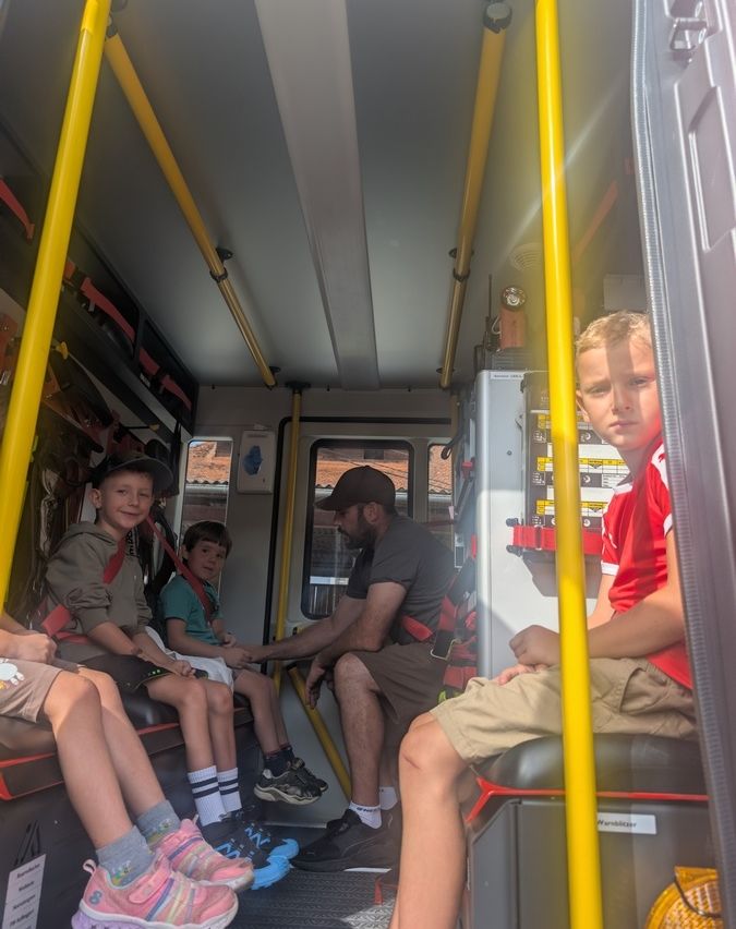 Vier Kinder und ein erwachsener Mann sitzen in einem Krankenwagen. Die Kinder tragen Freizeitkleidung und scheinen die Erwachsenen anzuschauen. Der Erwachsene trägt einen Hut und scheint mit den Kindern zu sprechen.