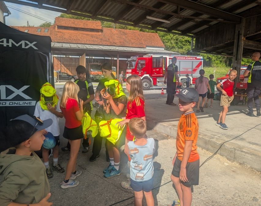 Eine Gruppe von Kindern, die Sicherheitswesten tragen, versammelt sich in einem überdachten Bereich, möglicherweise für eine Sicherheitsdemonstration. Ein Feuerwehrauto ist in der Nähe geparkt.
