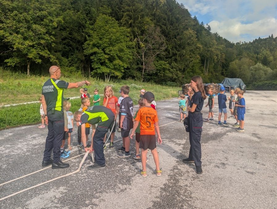 Eine Gruppe von Kindern und Erwachsenen versammelt sich auf einer Straße, wobei zwei Erwachsene in Uniformen den Kindern etwas demonstrieren. Die Kinder stehen in einer Reihe, und einige tragen Turnschuhe. Die Erwachsenen stehen auf beiden Seiten der Kinder. In der Ferne gibt es Bäume und einen Haufen Holz.