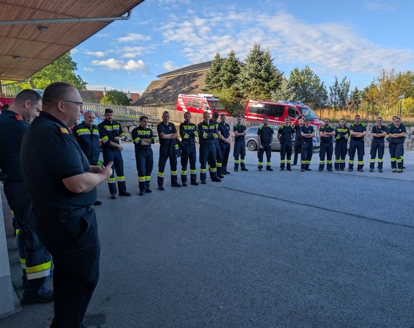 Eine Gruppe von Feuerwehrleuten in Uniform steht in einer Reihe, wobei ein Mann vor ihnen spricht. Sie sind draußen, mit einem roten Feuerwehrwagen, der hinter ihnen geparkt ist.