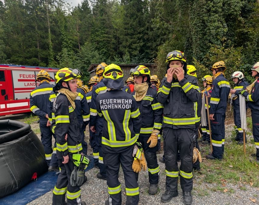 Eine Gruppe Feuerwehrleute in voller Ausrüstung, einschließlich Helmen und gelben reflektierenden Streifen, versammelt sich in einer Waldumgebung. Einige tragen Masken. Ein Feuerwehrauto ist im Hintergrund sichtbar.