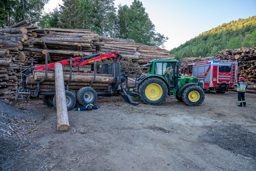 A green tractor with a red log loader is parked next to a pile of logs. A fire truck is parked behind it.