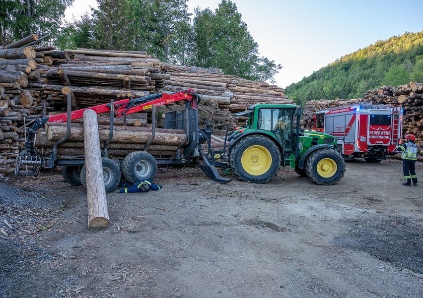 Ein grüner Traktor mit einem roten Holzlader steht neben einem Holzstapel. Dahinter ist ein Feuerwehrauto geparkt.
