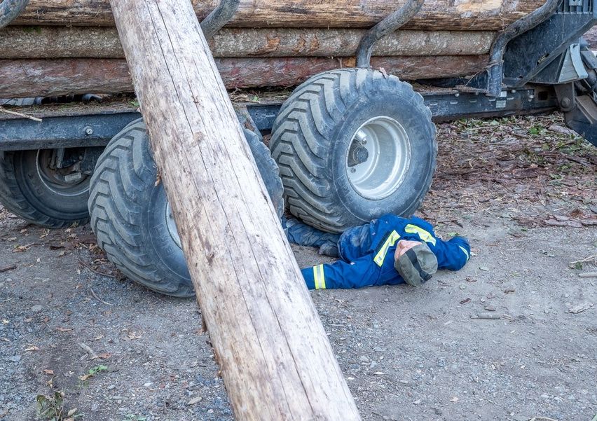 A worker lies on the ground near a truck loaded with logs, possibly injured.