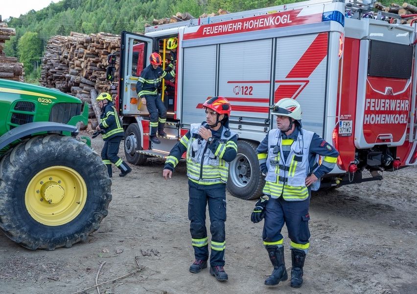 Feuerwehrleute in Schutzkleidung stehen vor einem Feuerwehrwagen, einer klettert in das Fahrzeug. Ein Traktor und ein Stapel Holzscheite sind im Hintergrund.