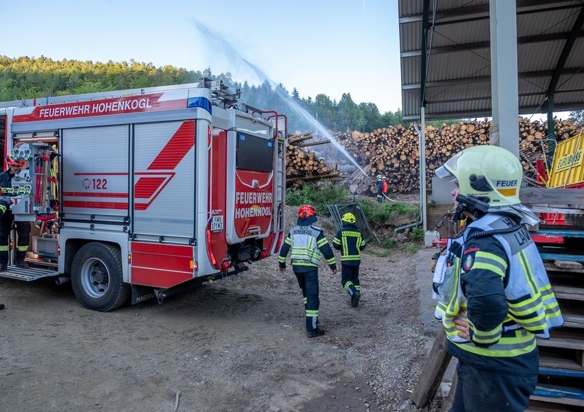 Firefighters in protective gear are walking towards a fire truck spraying water on a pile of logs. The scene is set in a forest area with a clear sky.