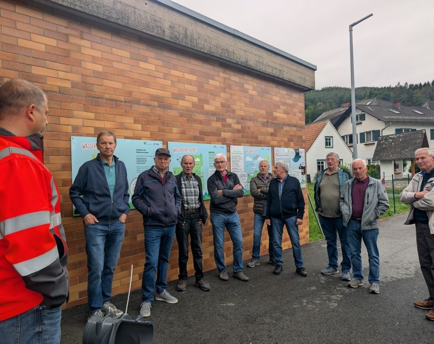 Eine Gruppe von Männern in Jacken und Jeans steht vor einer Ziegelmauer mit Plakaten, ein Mann in einer roten Jacke hört aufmerksam zu.