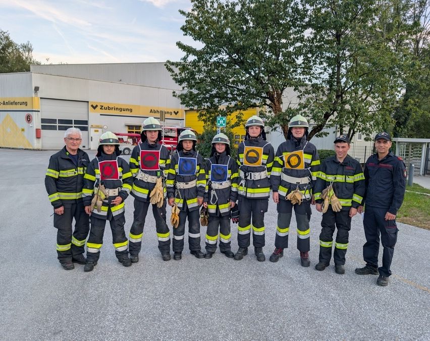 Eine Gruppe Feuerwehrleute in voller Ausrüstung steht vor einer Feuerwache mit einem Baum im Hintergrund.
