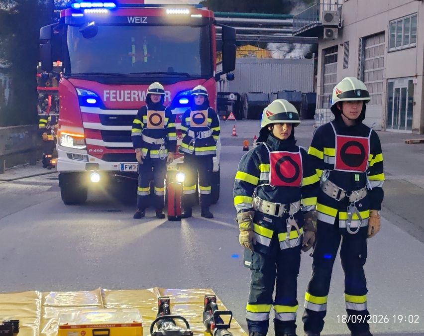 Drei Feuerwehrleute stehen vor einem roten Feuerwehrauto mit eingeschalteten Lichtern, in der Nähe eines Gebäudes, aus dem Rauch aufsteigt.
