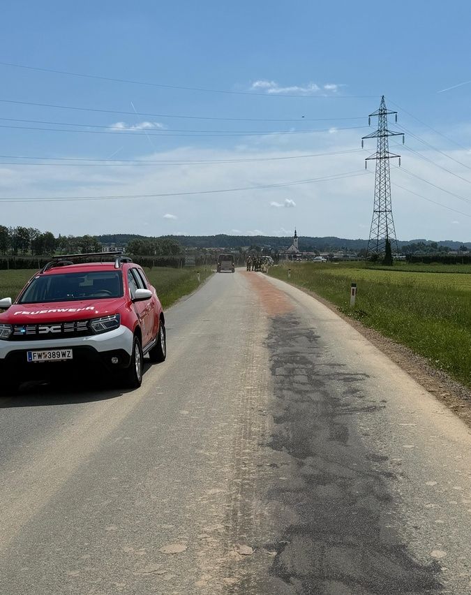 Ein roter SUV fährt auf einer Landstraße mit einem Dorf und einem Turm im Hintergrund.