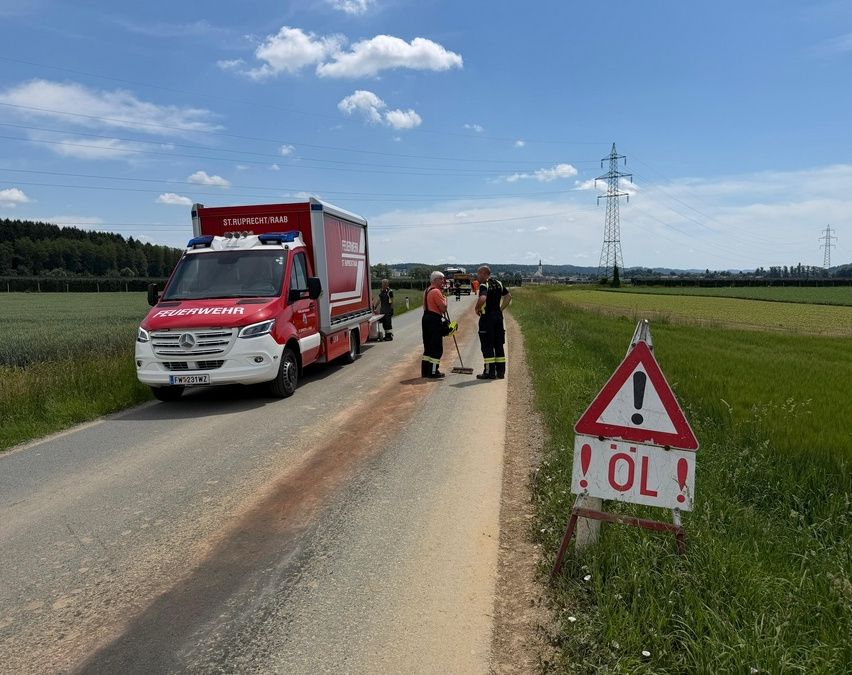 Ein Feuerwehrwagen ist auf der Straße. Zwei Personen reinigen die Straße mit einem Besen. Ein Schild warnt vor Ölverschmutzung.