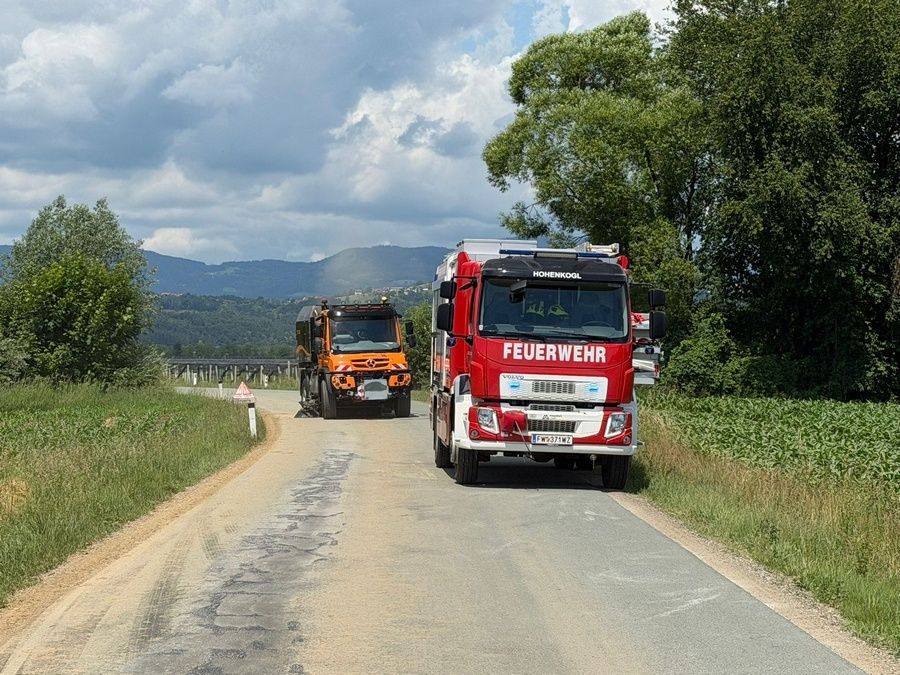 Zwei Feuerwehrfahrzeuge fahren auf einer Landstraße mit Gras und Bäumen auf beiden Seiten. Ein LKW ist rot mit dem Wort Feuerwehr auf der Front.