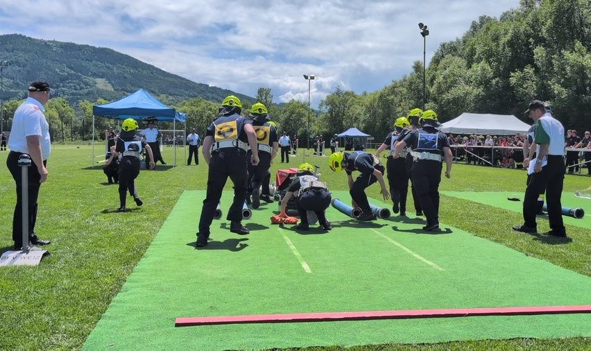 Feuerwehrleute in gelben Helmen üben auf einem grünen Feld, Zelte und Zuschauer im Hintergrund.