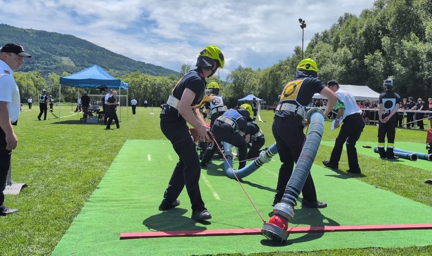 Eine Gruppe von Feuerwehrleuten übt eine Übung auf einem grünen Feld. Sie benutzen einen großen Schlauch und eine Schlauchtrommel. Das Gelände ist von grünem Gras und Zelten umgeben.