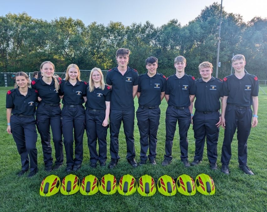 Eine Gruppe junger Menschen in Uniform posiert für ein Foto auf einem grünen Feld mit gelben Helmen davor.