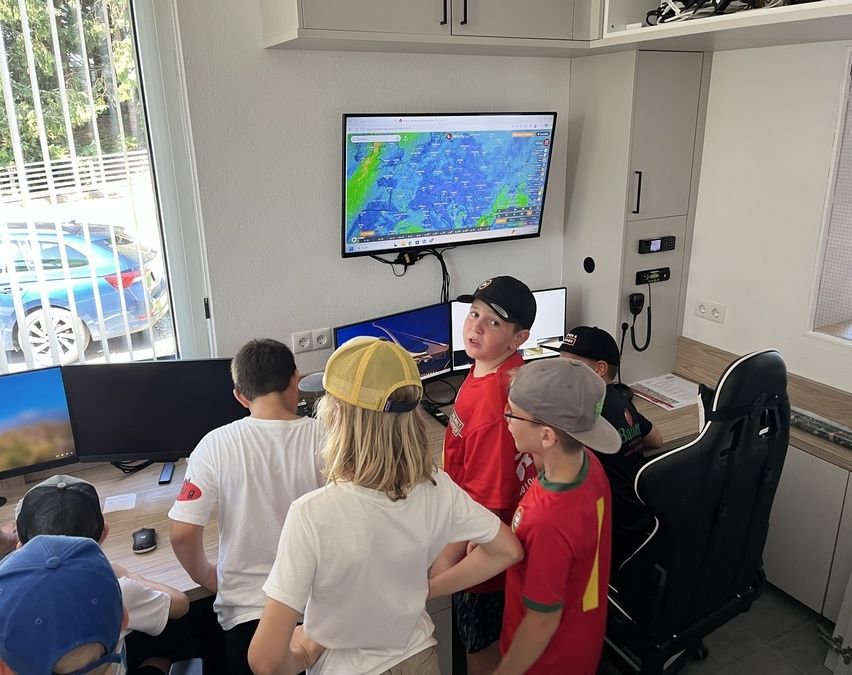 Mehrere Kinder sind in einem Raum und beobachten eine Wetterkarte auf einem an der Wand montierten Monitor. Jedes Kind trägt einen Hut und sie stehen um Computer herum.