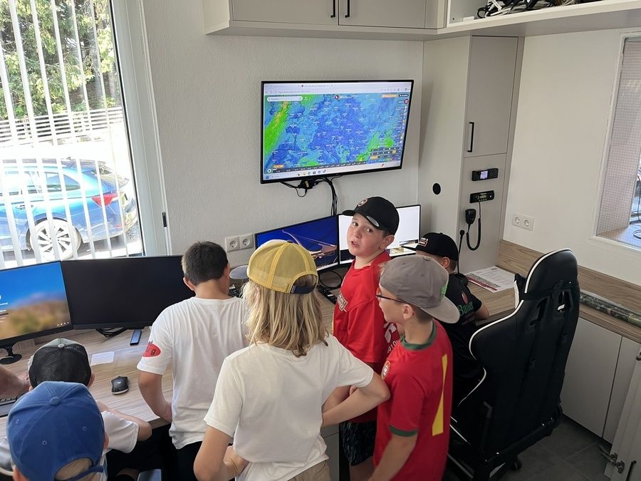 Mehrere Kinder sind in einem Raum und beobachten eine Wetterkarte auf einem an der Wand montierten Monitor. Jedes Kind trägt einen Hut und sie stehen um Computer herum.