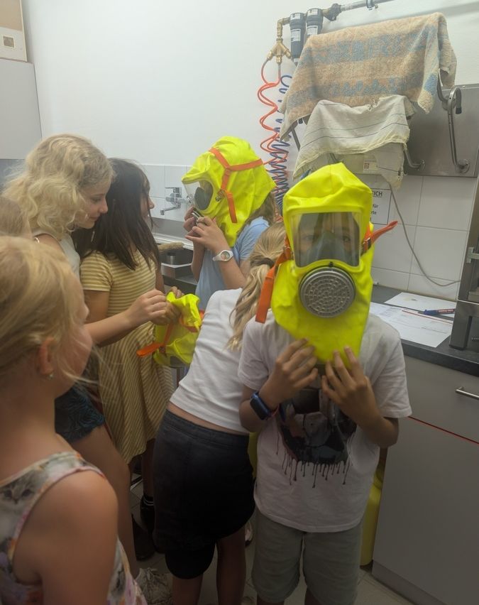 Mehrere Kinder in Schutzanzügen und Masken stehen in einem Labor, eines hält eine Maske vors Gesicht.