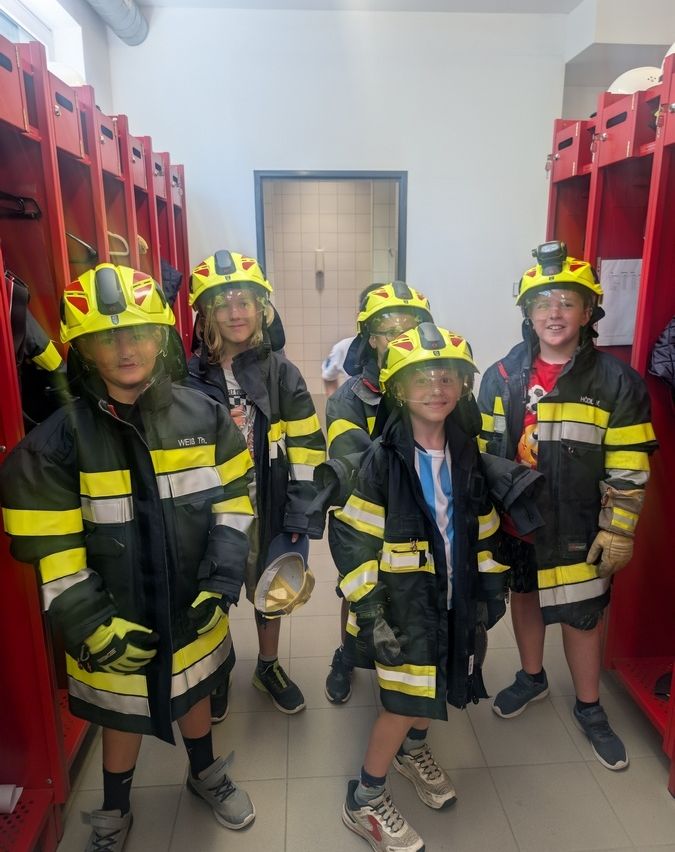 Mehrere Kinder in Feuerwehruniformen stehen in einem Raum und posieren für ein Foto. Sie haben Helme und Handschuhe an. Hinter ihnen sind rote Schließfächer zu sehen. Der Raum hat geflieste Böden.