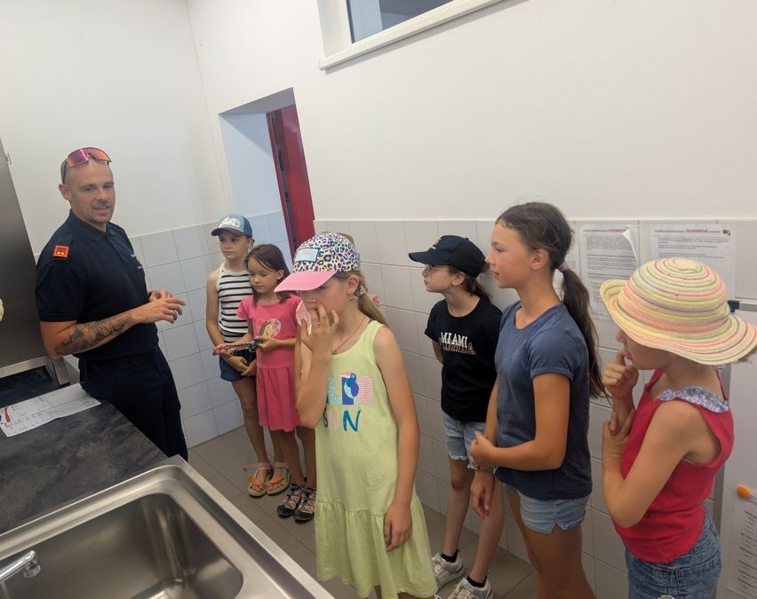 Ein Mann in Uniform spricht mit einer Gruppe junger Mädchen in einem Raum. Sie stehen vor einer weißen Wand, mit einem Waschbecken und einem Fenster in der Nähe.