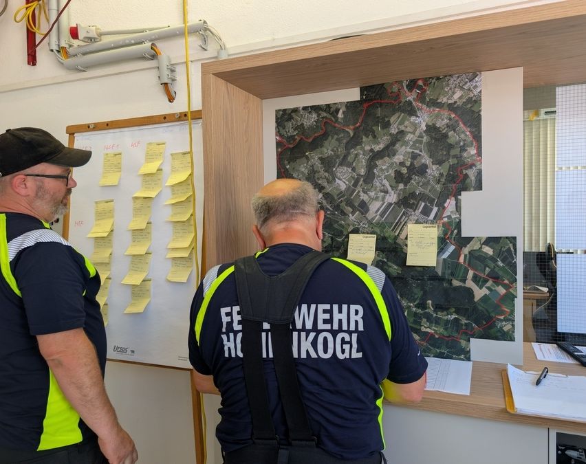 Zwei Feuerwehrleute überprüfen eine Karte mit roten Markierungen und Notizen auf einem Brett. Einer trägt einen Rucksack, und der andere schaut auf das Brett.