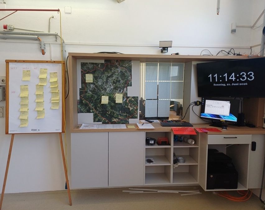 Ein Computeraufbau mit einem Monitor, der die Zeit anzeigt, einer Tastatur und verschiedenen Gegenständen auf einem Schreibtisch. Eine Tafel mit Haftnotizen und einer gerahmten Karte an der Wand.