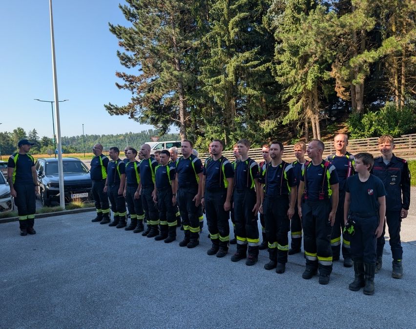 Eine Gruppe von Feuerwehrleuten steht in einer Reihe, in ihren Uniformen gekleidet, vor einem Pfosten und Bäumen.