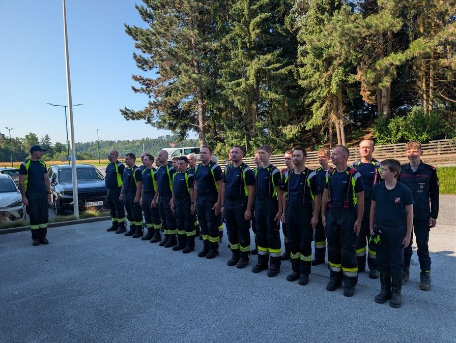 Eine Gruppe von Feuerwehrleuten steht in einer Reihe, in ihren Uniformen gekleidet, vor einem Pfosten und Bäumen.