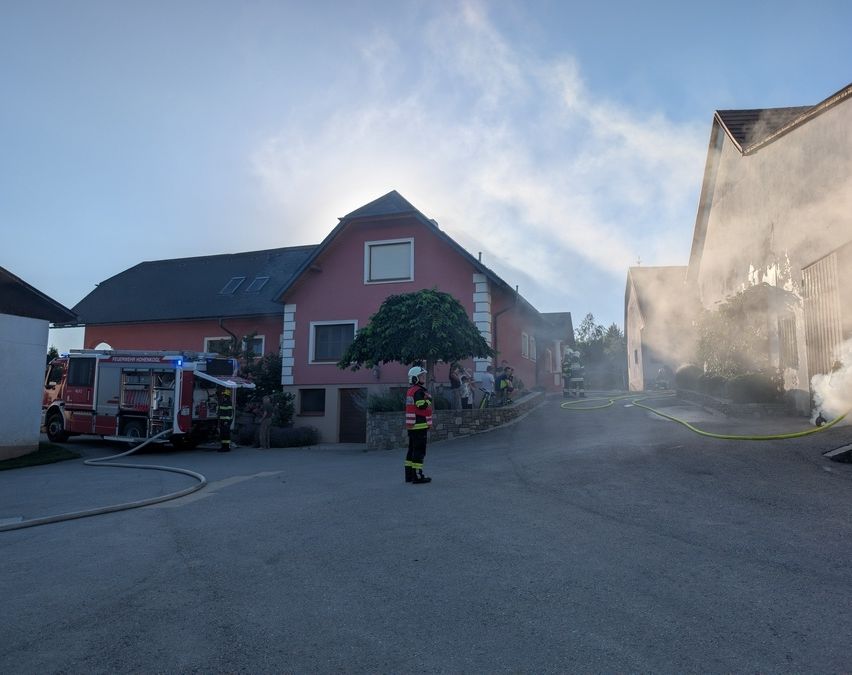 Ein Feuerwehrwagen steht auf der Straße mit Feuerwehrleuten vor einem roten Haus. Rauch steigt vom Dach auf.