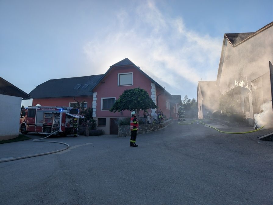 Ein Feuerwehrwagen steht auf der Straße mit Feuerwehrleuten vor einem roten Haus. Rauch steigt vom Dach auf.