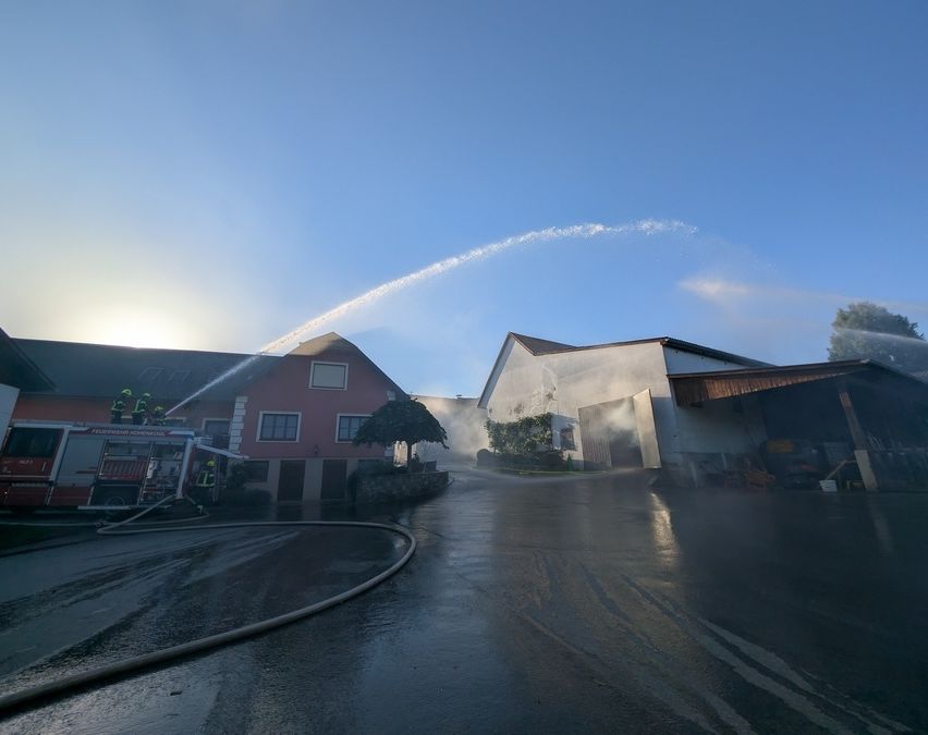 Ein Feuerwehrauto spritzt Wasser auf ein Haus an einem sonnigen Tag, wobei Feuerwehrleute auf dem Dach stehen und ein Baum im Vordergrund steht.