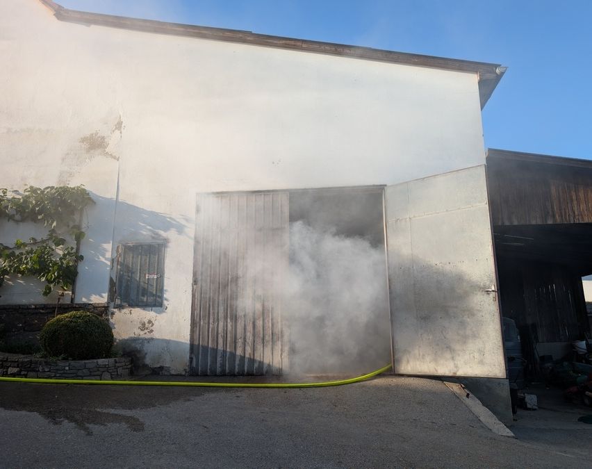 Rauch dringt aus einer großen Metalltür eines weißen Gebäudes. Ein gelber Schlauch liegt auf dem Boden.