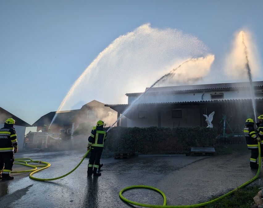 Feuerwehrleute sprühen Wasser auf ein Gebäude in Notlage mit einem Feuerwehrwagen im Hintergrund.