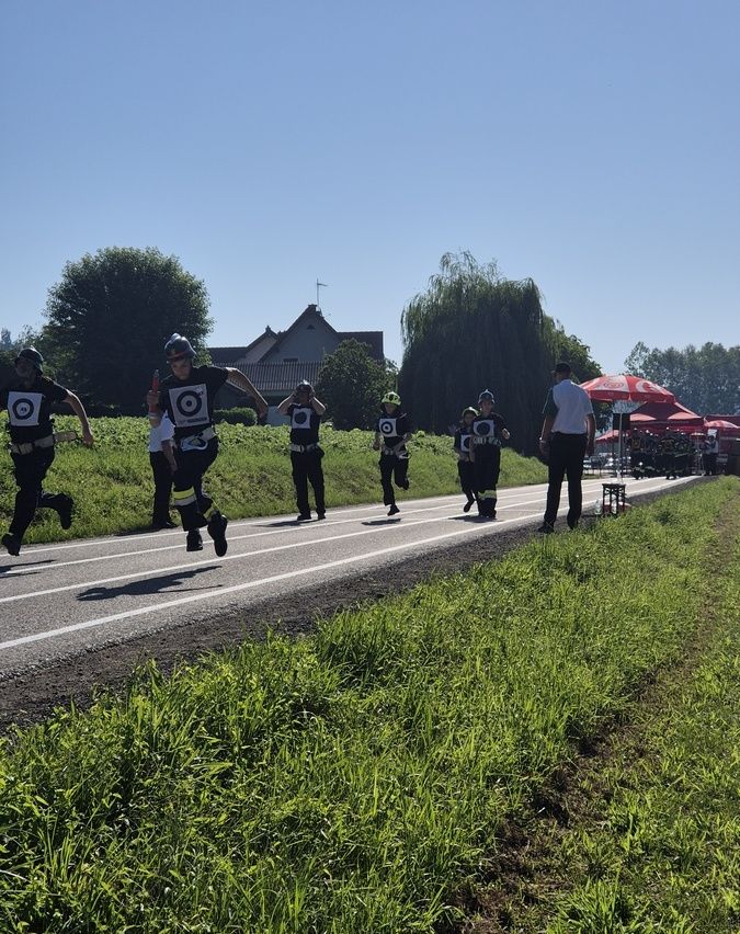 Eine Gruppe von Menschen in Helmen und Schutzausrüstung läuft auf einer Laufbahn. Ein Mann steht hinter ihnen, und ein roter Regenschirm ist in der Nähe. Bäume und Häuser sind im Hintergrund.