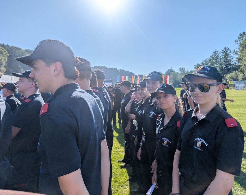 Eine Gruppe von Feuerwehrleuten in schwarzen Uniformen steht in einer Reihe auf einem Feld, einige tragen Mützen und Sonnenbrillen. Hinter ihnen stehen weitere Personen, möglicherweise weitere Feuerwehrleute, und ein Grasbereich mit Bäumen.