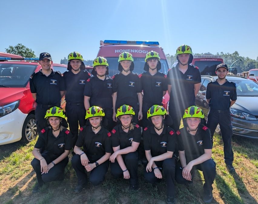 Eine Gruppe Feuerwehrleute in grünen Helmen und schwarzen Uniformen posiert für ein Foto vor Rettungsfahrzeugen.