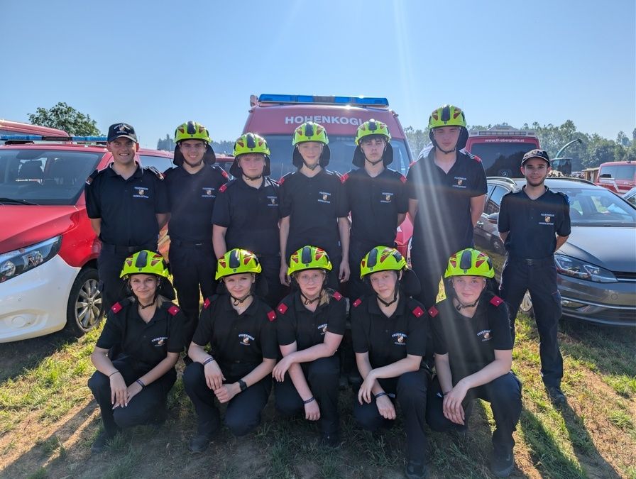 Eine Gruppe Feuerwehrleute in grünen Helmen und schwarzen Uniformen posiert für ein Foto vor Rettungsfahrzeugen.