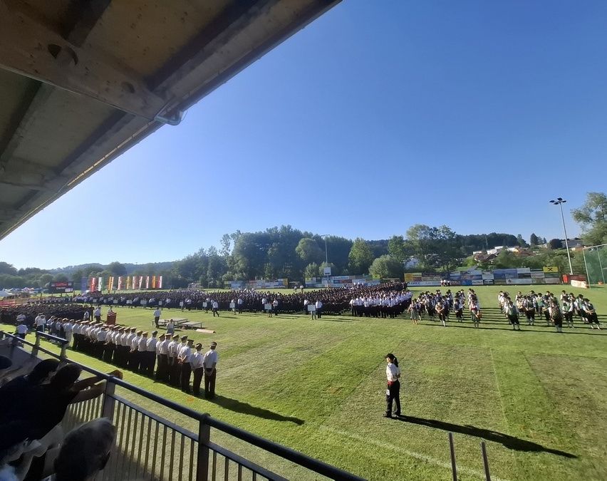 Eine große Gruppe von Menschen in Uniform steht auf einem Feld, einige in Formation. Zuschauer beobachten vom Balkon. Bäume und Berge sind in der Ferne.
