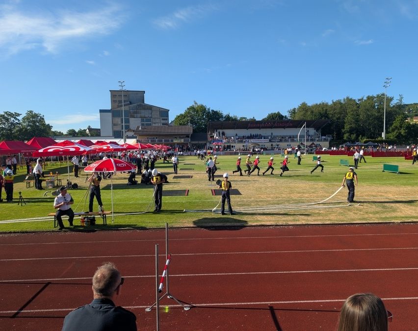 Eine Gruppe von Menschen nimmt an einer Leichtathletikveranstaltung im Freien teil. Mehrere Athleten laufen auf der Laufbahn, während andere sich auf ihren Einsatz vorbereiten. Rote Schirme und Bänke sind auf dem Feld aufgestellt.