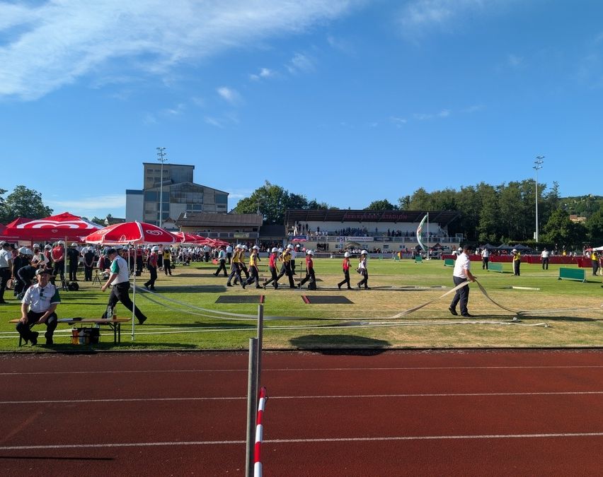 Menschen in einem Stadion, einige halten eine lange Schnur. Zuschauer stehen an den Seitenlinien. Ein Athlet läuft auf der Laufbahn.