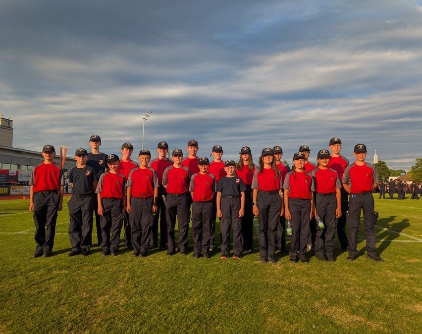 Eine Gruppe junger Menschen in roten und schwarzen Uniformen posiert für ein Foto auf einem Feld. Der Himmel ist bewölkt.
