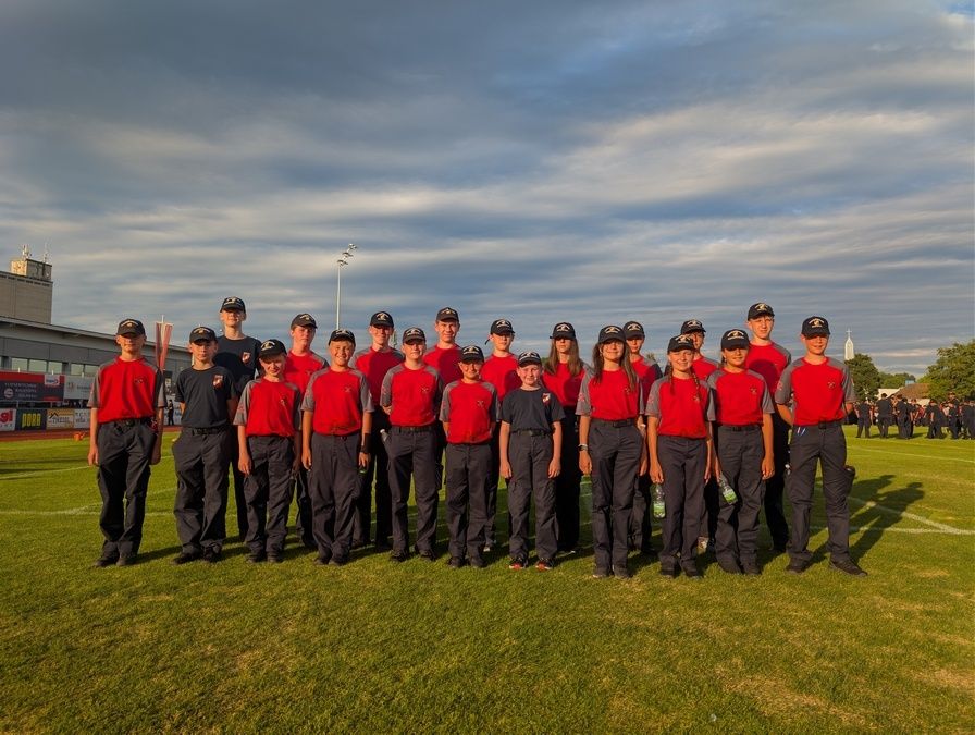 Eine Gruppe junger Menschen in roten und schwarzen Uniformen posiert für ein Foto auf einem Feld. Der Himmel ist bewölkt.