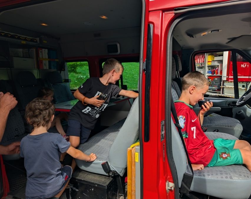 Mehrere Kinder befinden sich in einem roten Feuerwehrauto. Ein Junge hält ein Walkie-Talkie und scheint zu sprechen. Ein anderer Junge sitzt auf einem Sitz und hält ein Mikrofon.