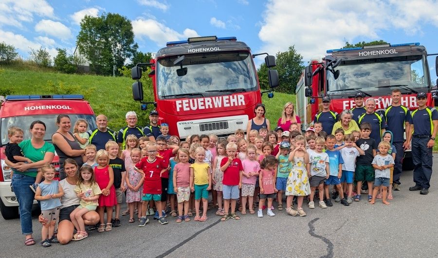 Eine Gruppe von Kindern und Erwachsenen posiert vor zwei Feuerwehrwagen, einem mit der Aufschrift 'Feuerwehr' und dem anderen 'Hohenkohl', unter einem blauen Himmel mit einigen Wolken und grünem Gras im Hintergrund.