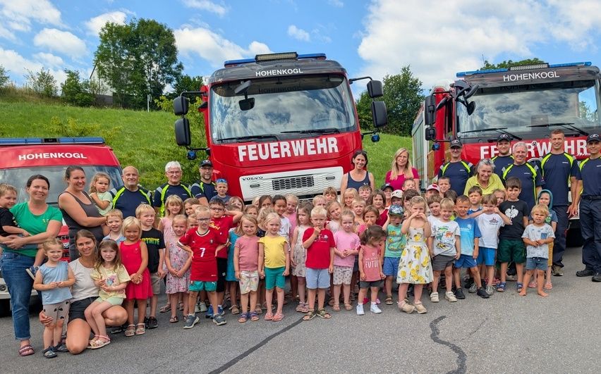 Eine Gruppe von Kindern und Erwachsenen posiert vor zwei Feuerwehrwagen, einem mit der Aufschrift 'Feuerwehr' und dem anderen 'Hohenkohl', unter einem blauen Himmel mit einigen Wolken und grünem Gras im Hintergrund.
