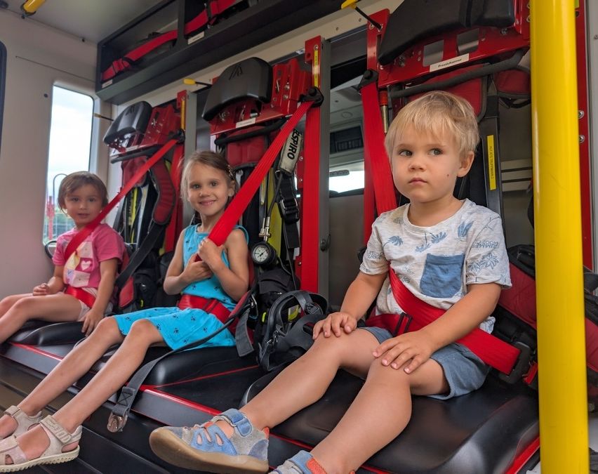 Drei Kinder, zwei Mädchen und ein Junge, sitzen in Sicherheitsgurten in einem roten Notfallfahrzeug, möglicherweise einem Krankenwagen, und schauen geradeaus.