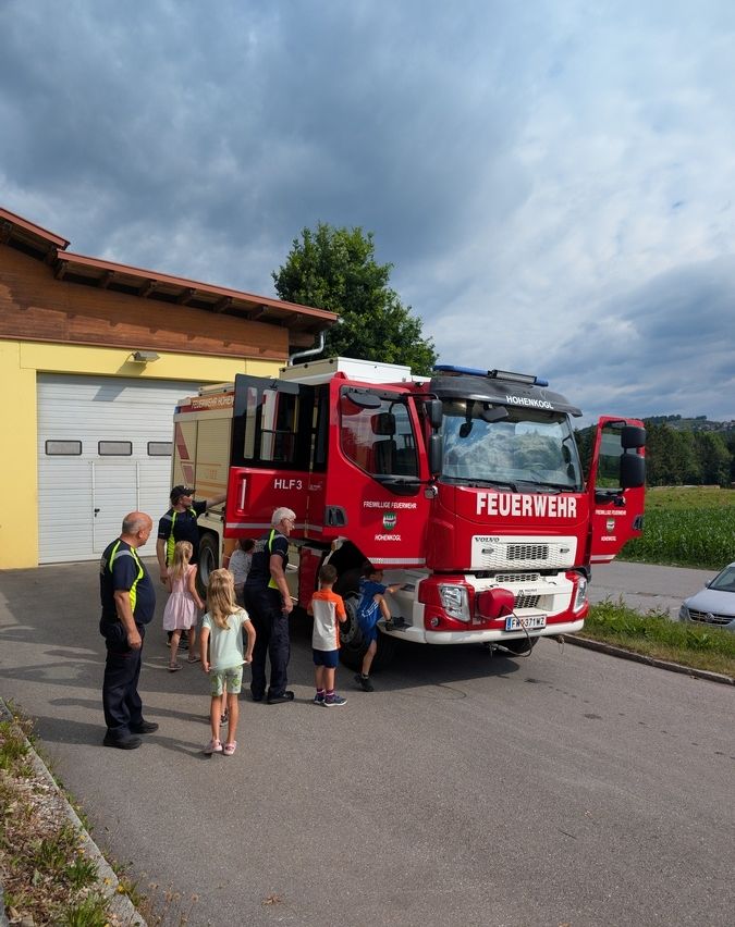 Ein Feuerwehrfahrzeug ist vor einem Gebäude geparkt, an dem mehrere Kinder und Erwachsene es betrachten.